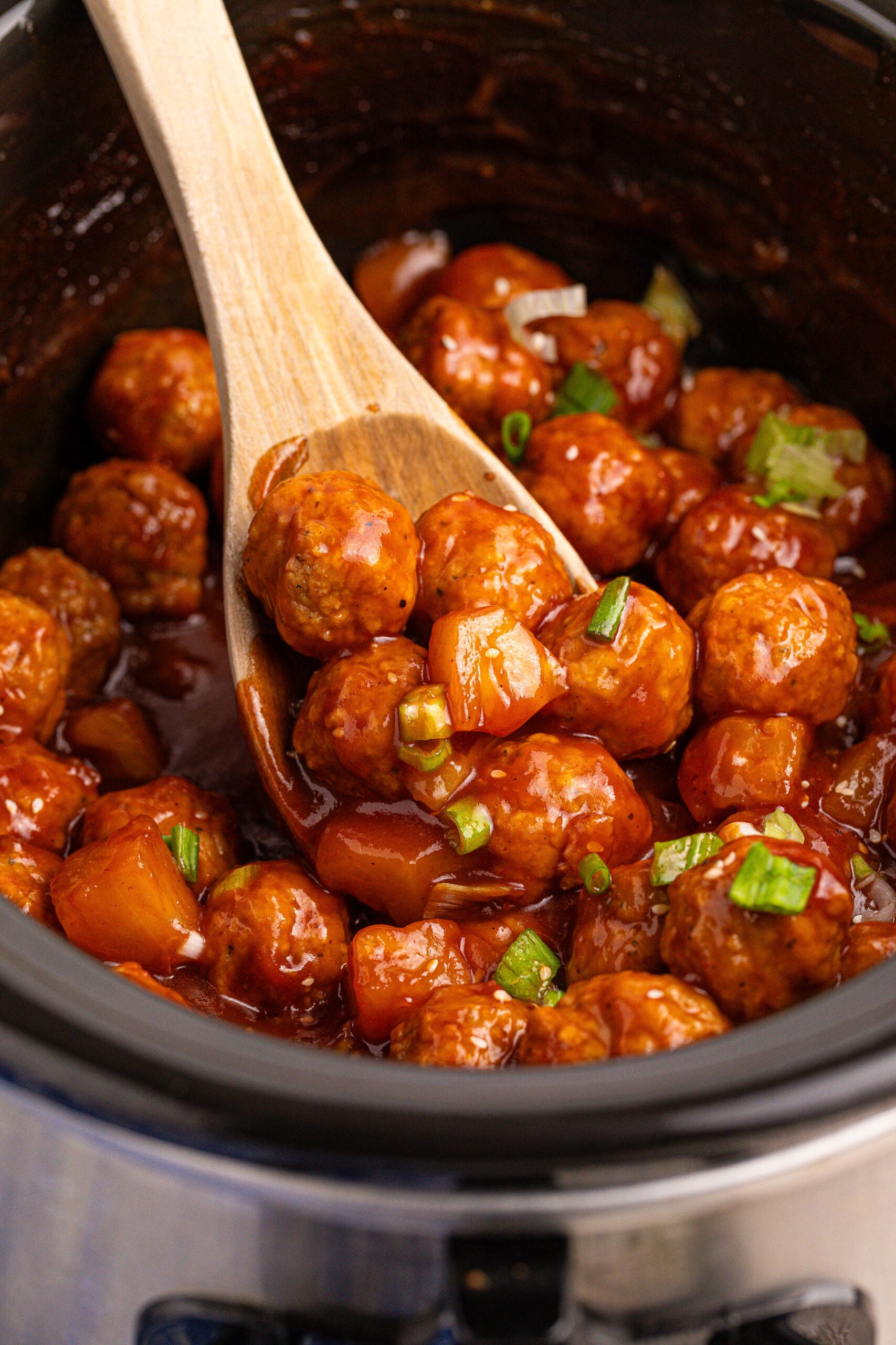 Slow Cooker 3 Ingredient Hawaiian Meatballs in a black crock pot with a wooden spoon lifting up a few 