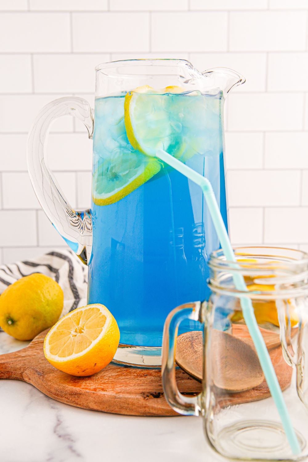 Blue Raspberry Lemonade in a pitcher and an empty glass with a straw sitting in the front of the pitcher