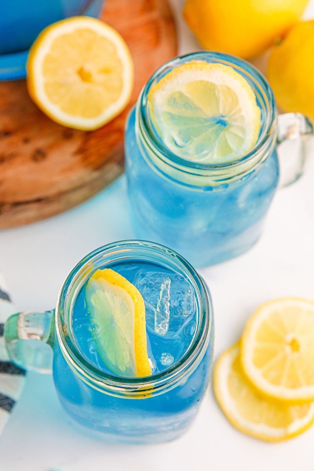 two glasses wit hBlue Raspberry Lemonade in them and lemons and a wooden cutting board behind it on table