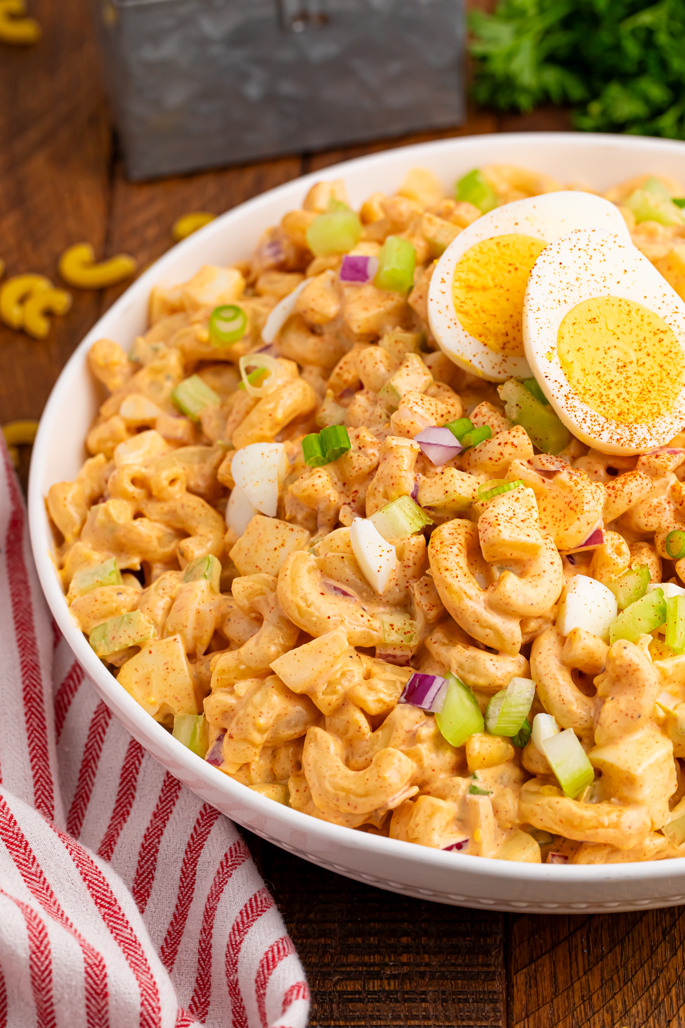 Deviled Egg Pasta Salad in a bowl sitting on wooden table with herbs around it and a red and white striped towel on table