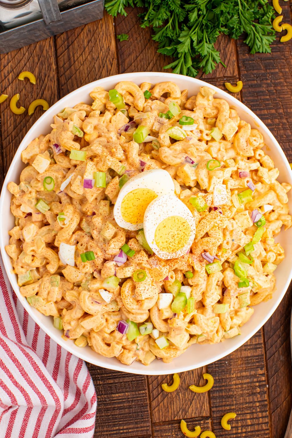 Deviled Egg Pasta Salad in a bowl sitting on wooden table with herbs around it and a red and white striped towel on table