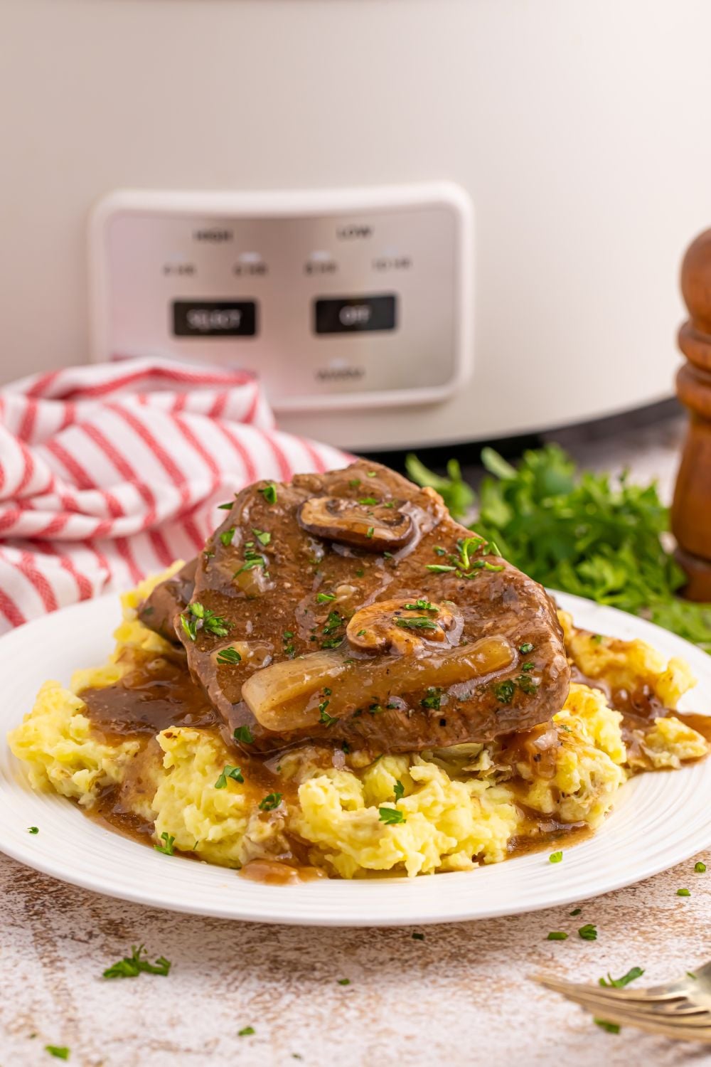 Slow Cooker Round Steak and Gravy on a plate with mashed potatoes under it. Then a red and white towel to the side with slow cooker in the background of picture