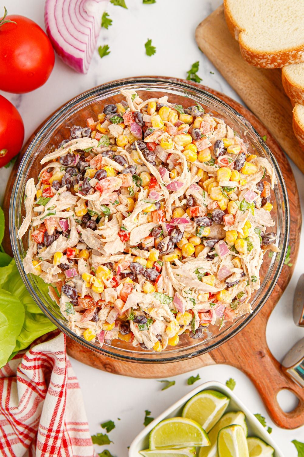 overhead of bowl with chicken salad in it and onions, tomatoes, limes and bread all around the bowl