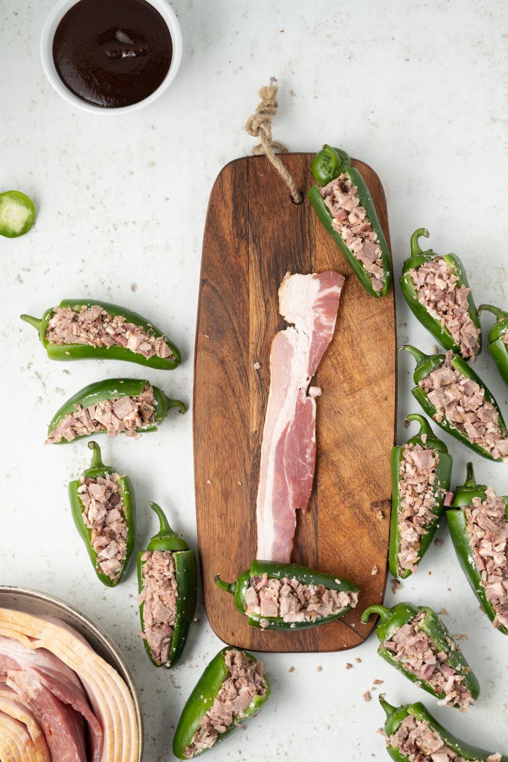 slice of raw bacon on cutting board with stuffed jalapenos sitting around it on table