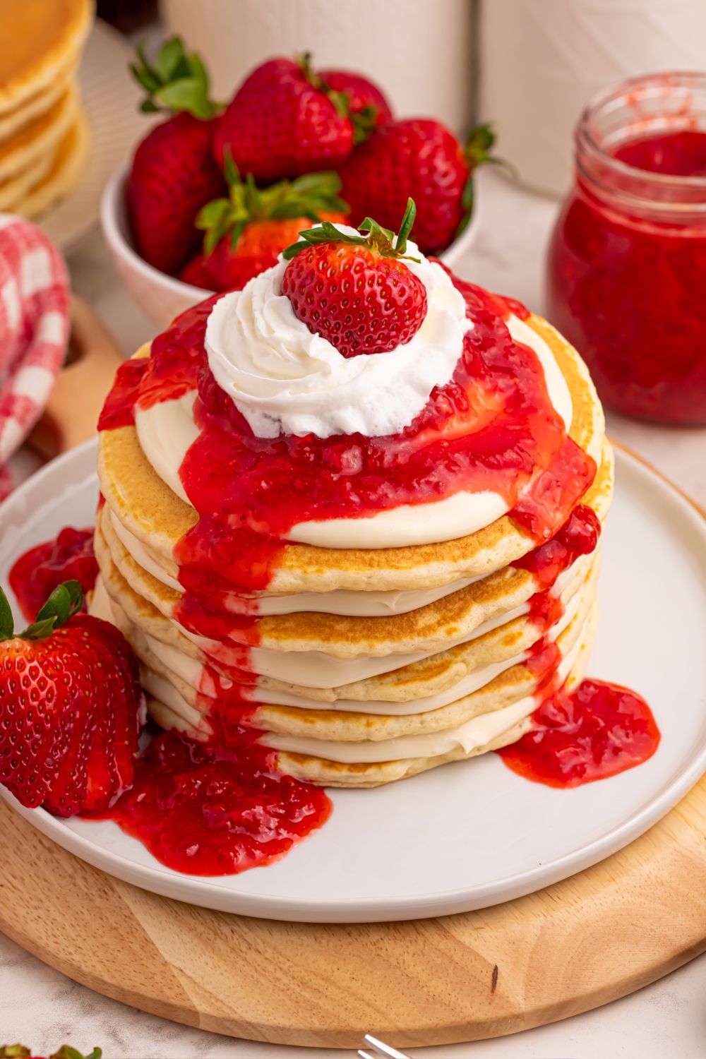 Strawberry Cheesecake Pancakes stacked up on plate and topped with whipped cream and a strawberry on top, with a bowl of berries behind it on table