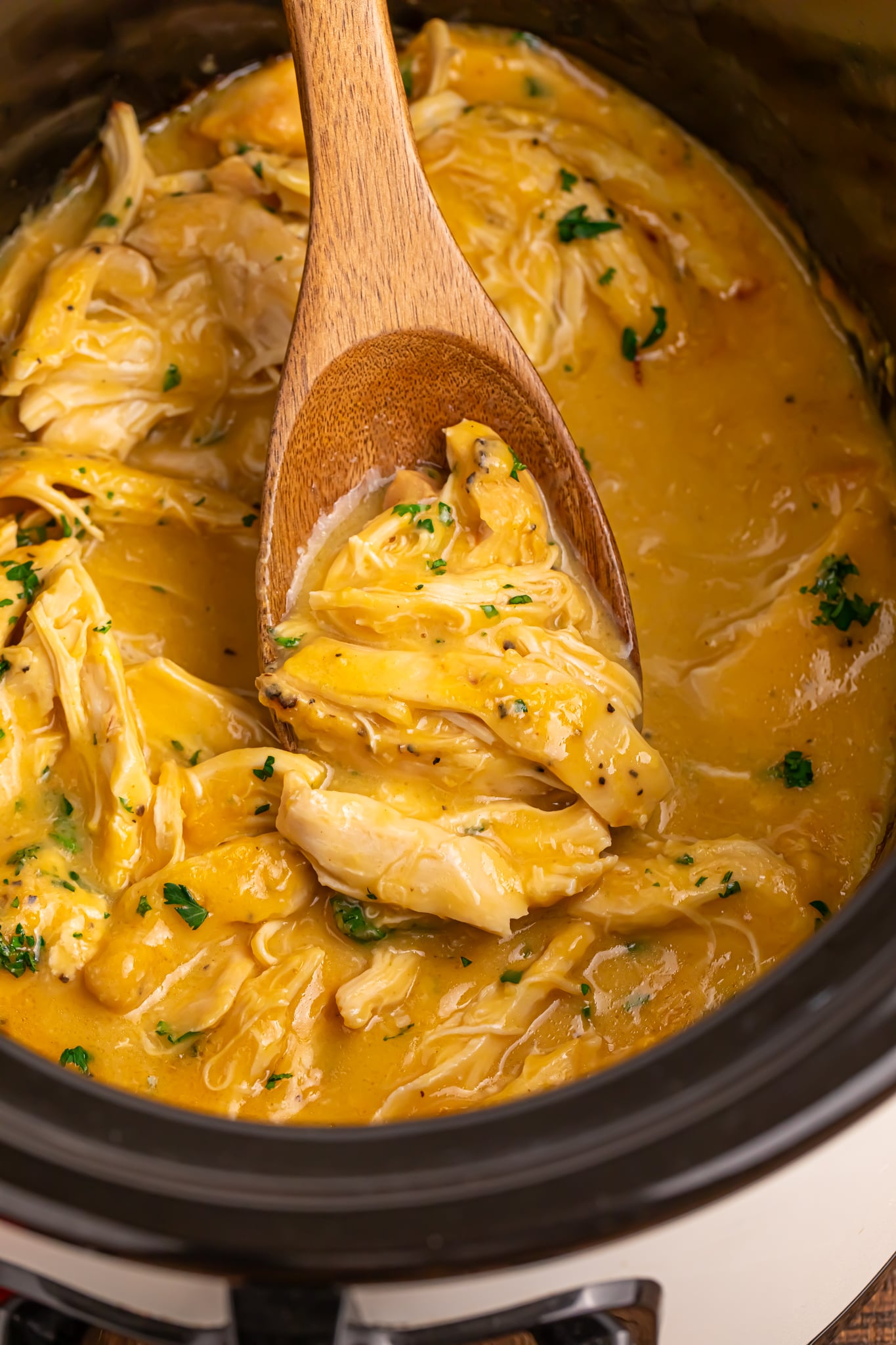 overhead look of crockpot with chicken and gravy in it with a wooden spoon with some meat and gravy on it 