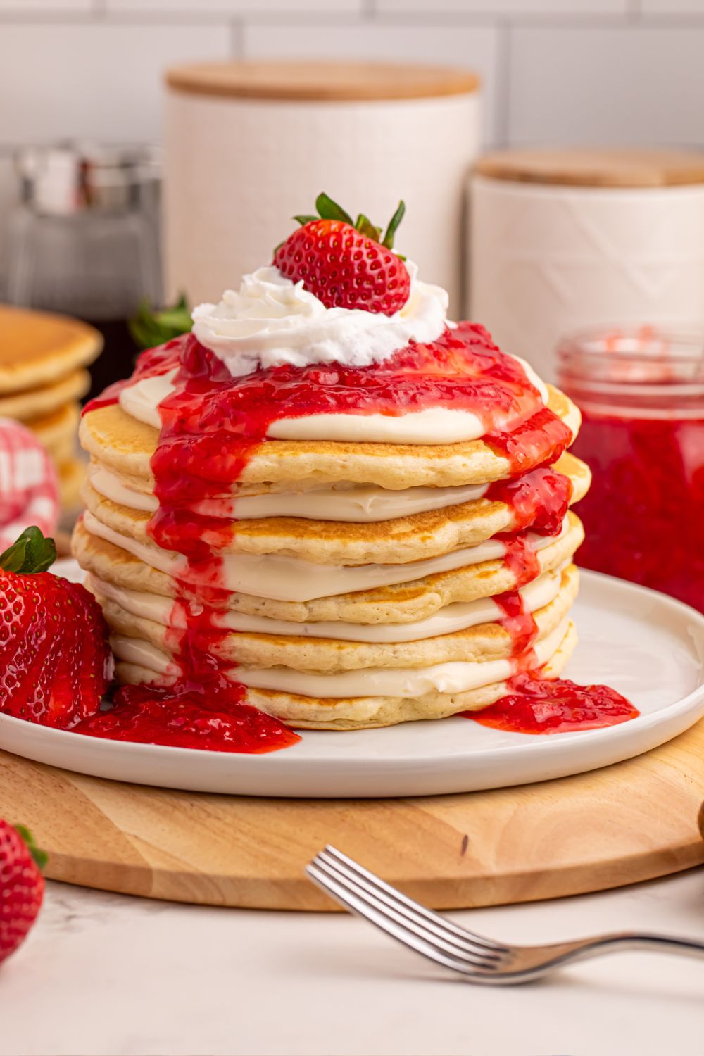Strawberry Cheesecake Pancakes sitting on plate on the table with sugar and flour containers in the background, a fork in the front, and strawberries