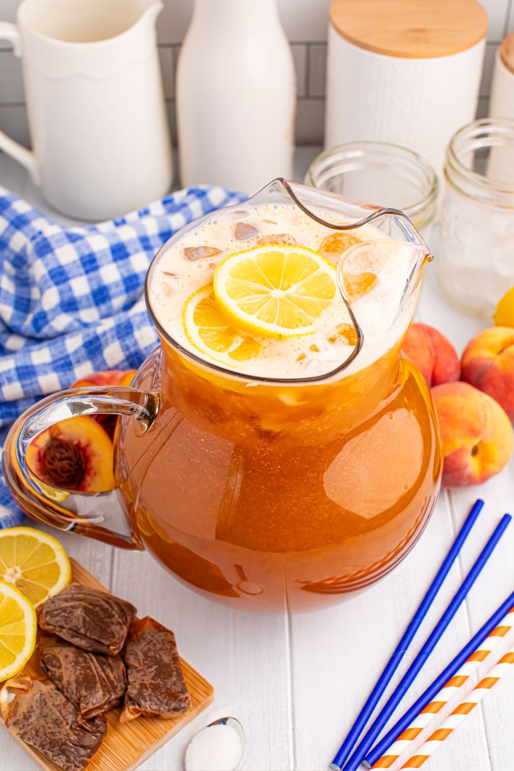 peach tea on the table with wet tea bags, slices of fruit, straws, sugar, and a blue checked towel around the container