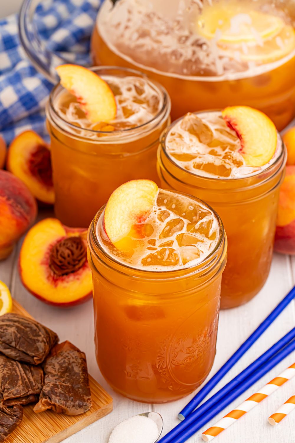 Peach Sweet Tea in a pitcher on table with 3 glasses in front of it, with tea bags, sliced peaches and more around it