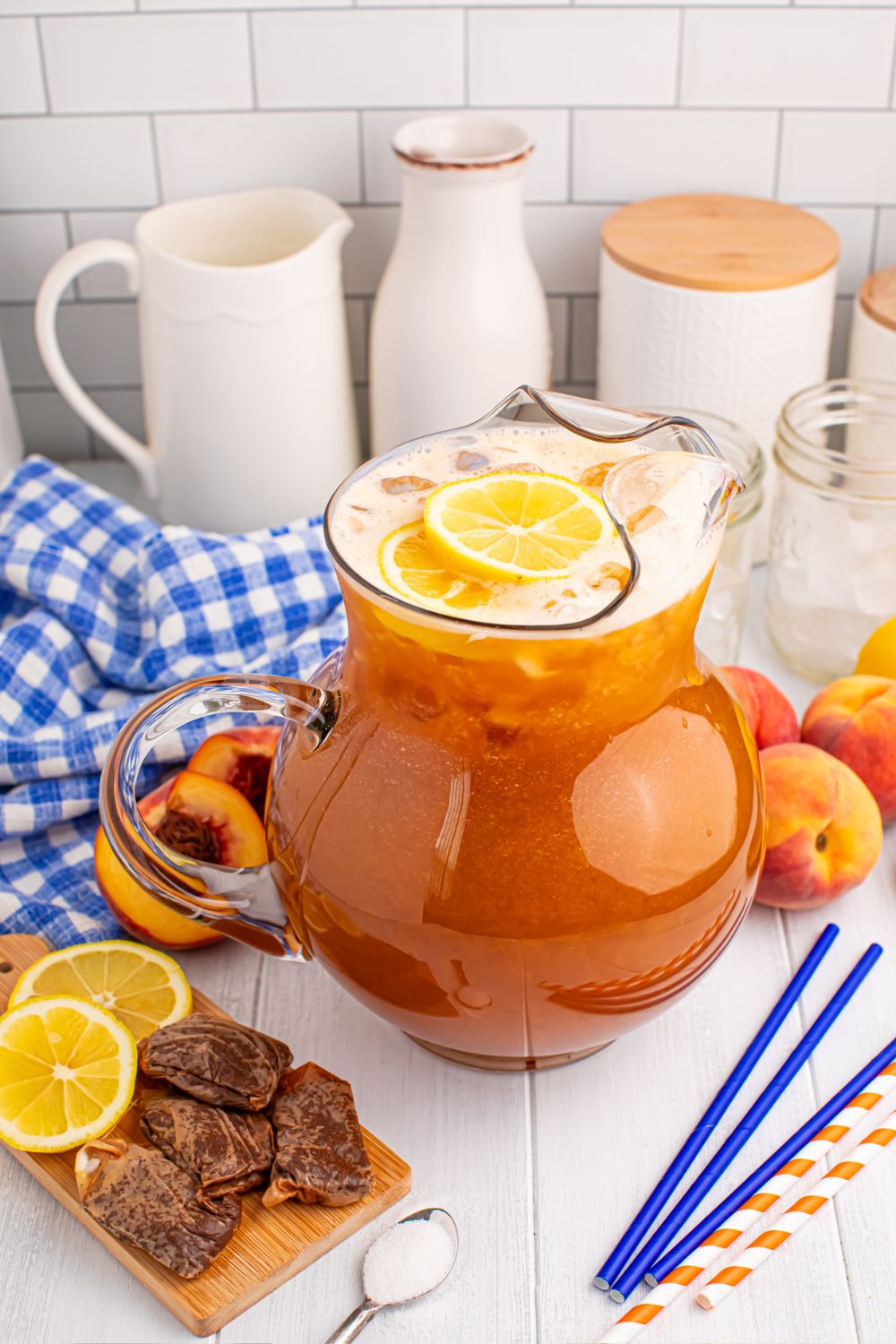 large pitcher of peach sweet tea on table with straws, sugar, lemon slices and more around the pitcher