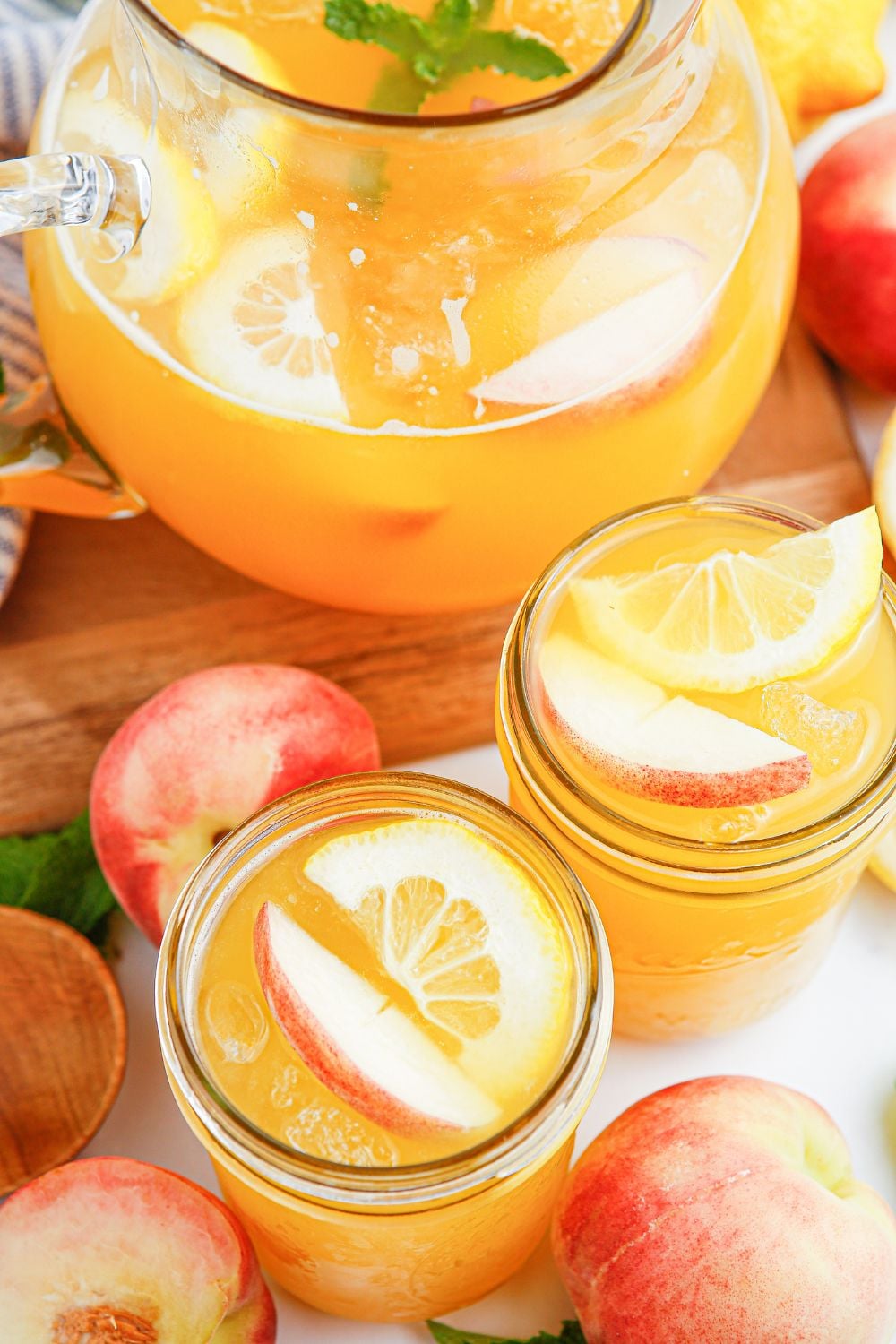 large pitcher with peach lemonade on table. Mason jars full of lemonade and fresh peaches scattered on table