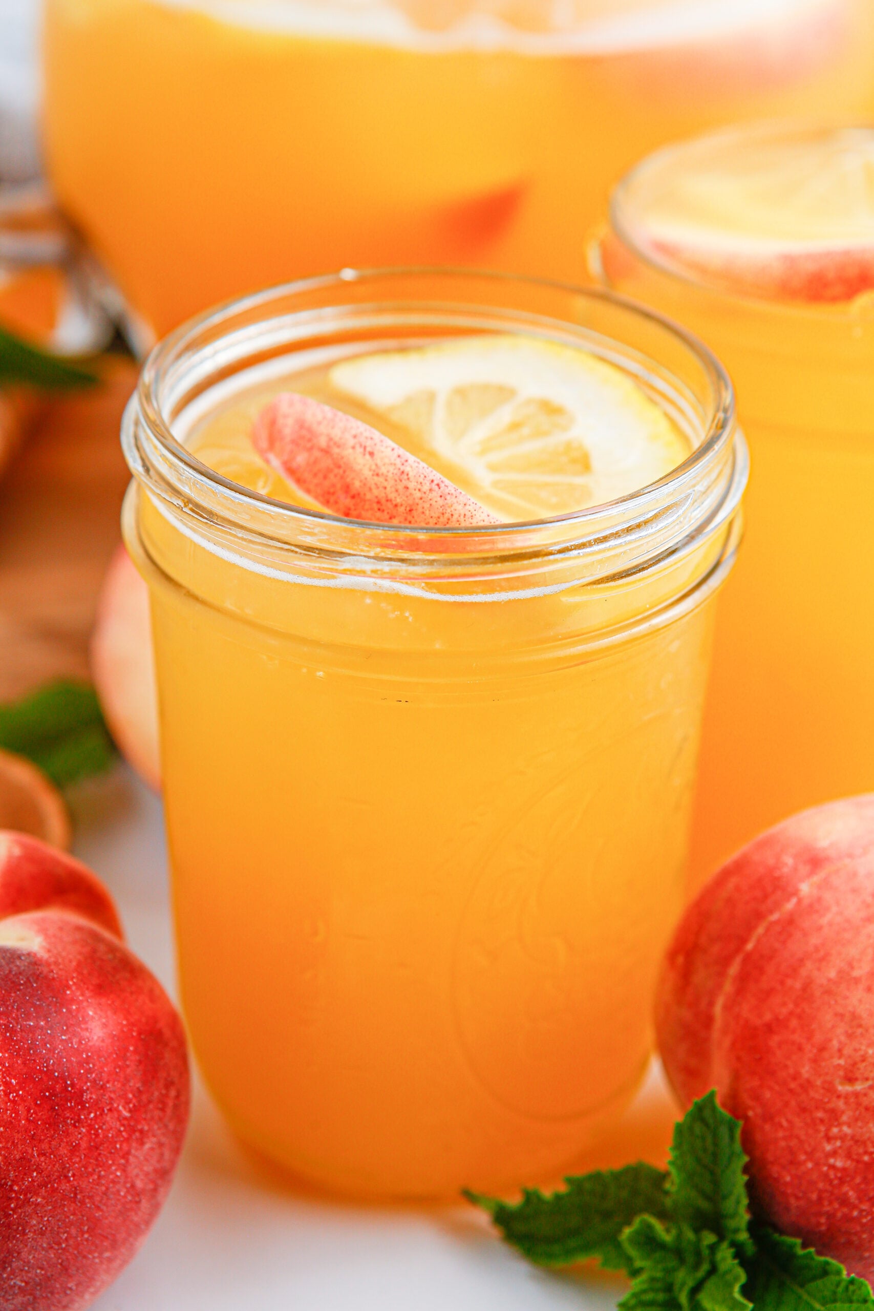 Peach Lemonade in a mason jar glass garnished with peach slice and orange slice. Fresh peaches around the glass