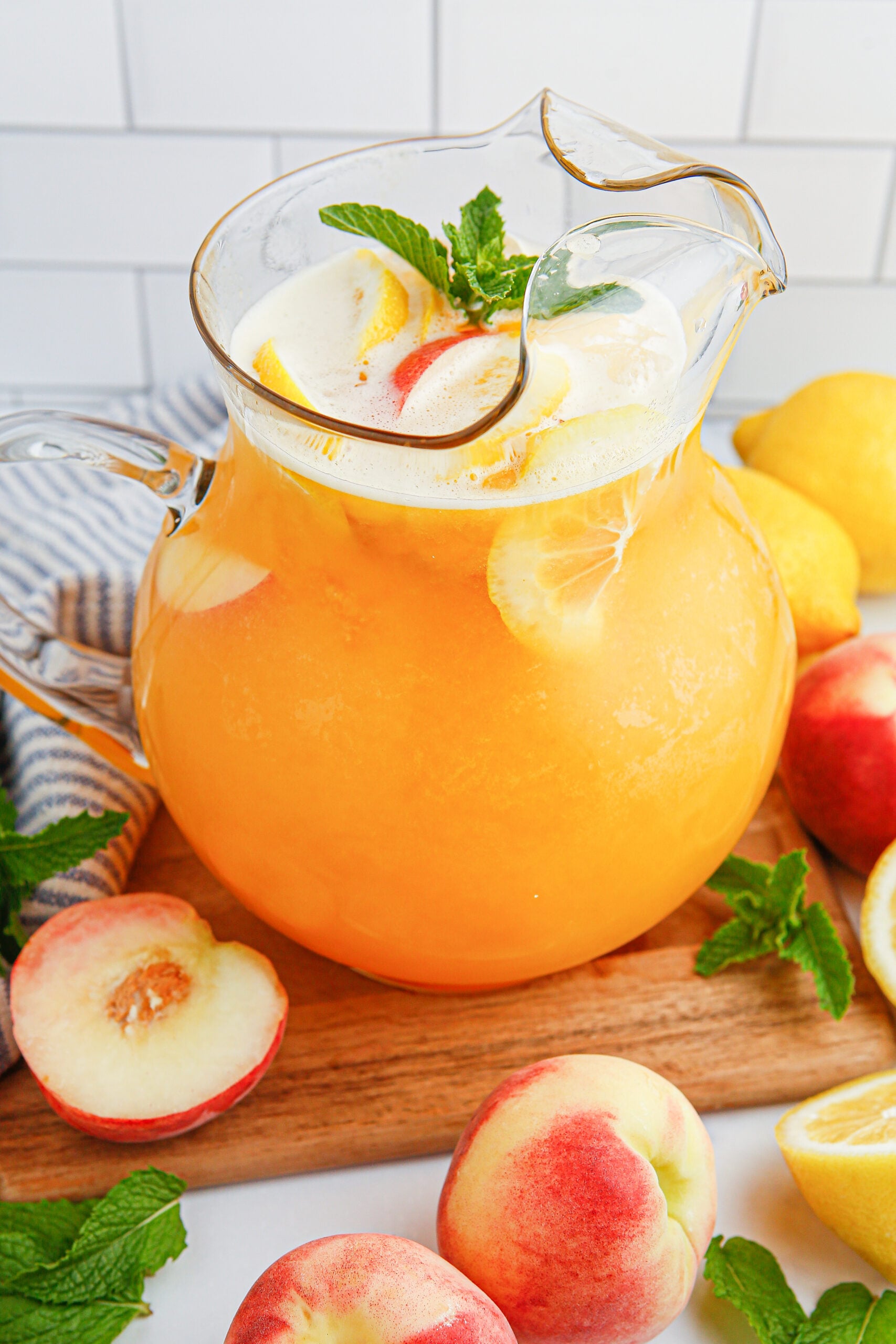 Peach Lemonade in a clear pitcher garnished with peach slice and orange slices. Fresh peaches around the glass