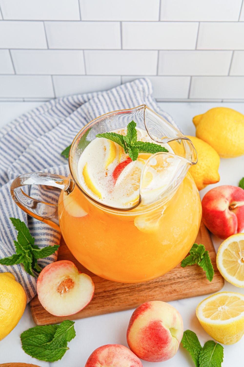 Peach Lemonade in a pitcher siting on a cutting board with peaches and lemon around it on table