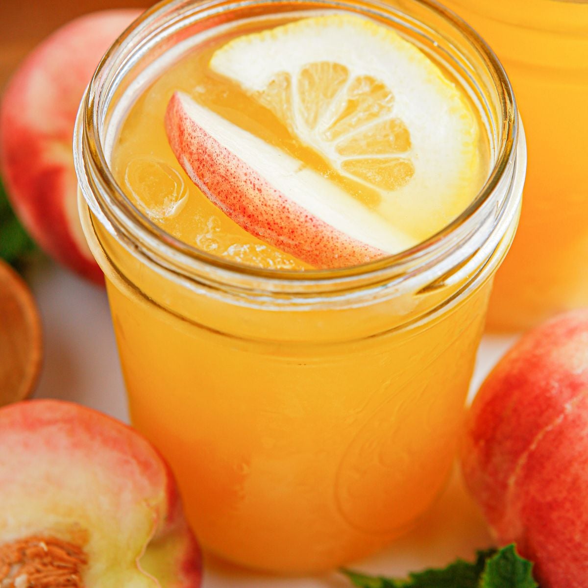 Peach Lemonade in a mason jar garnished with peach slice and orange slice. Fresh peaches around the glass