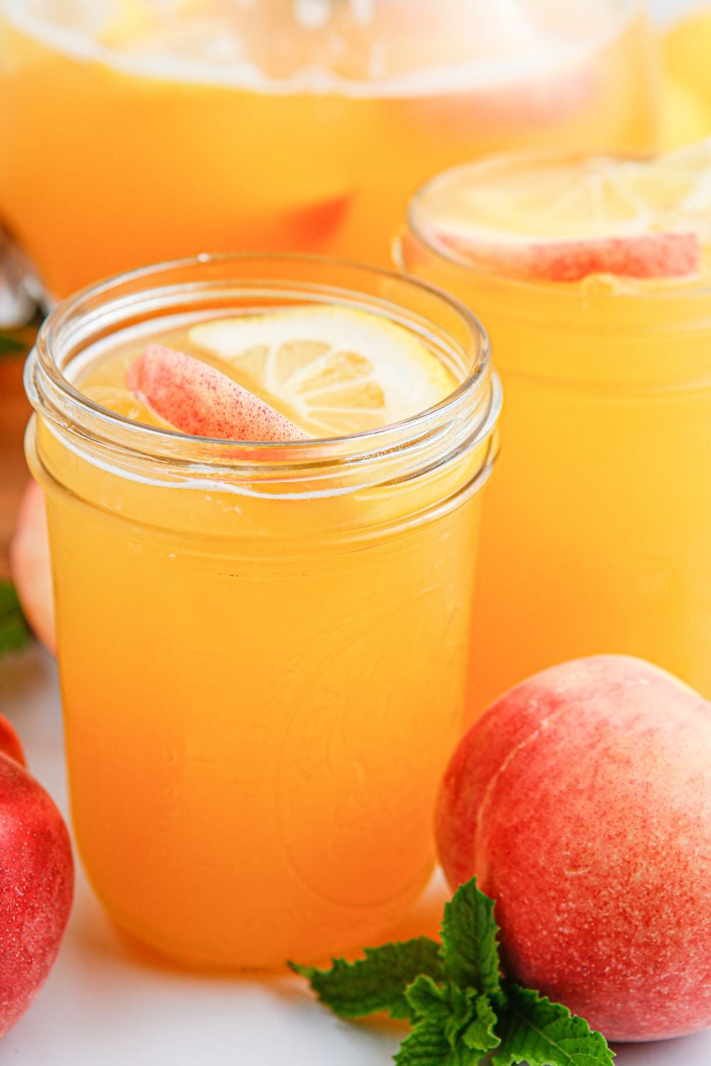 Peach Lemonade in mason jars sitting on table with a whole peach sitting to the side of the glasses