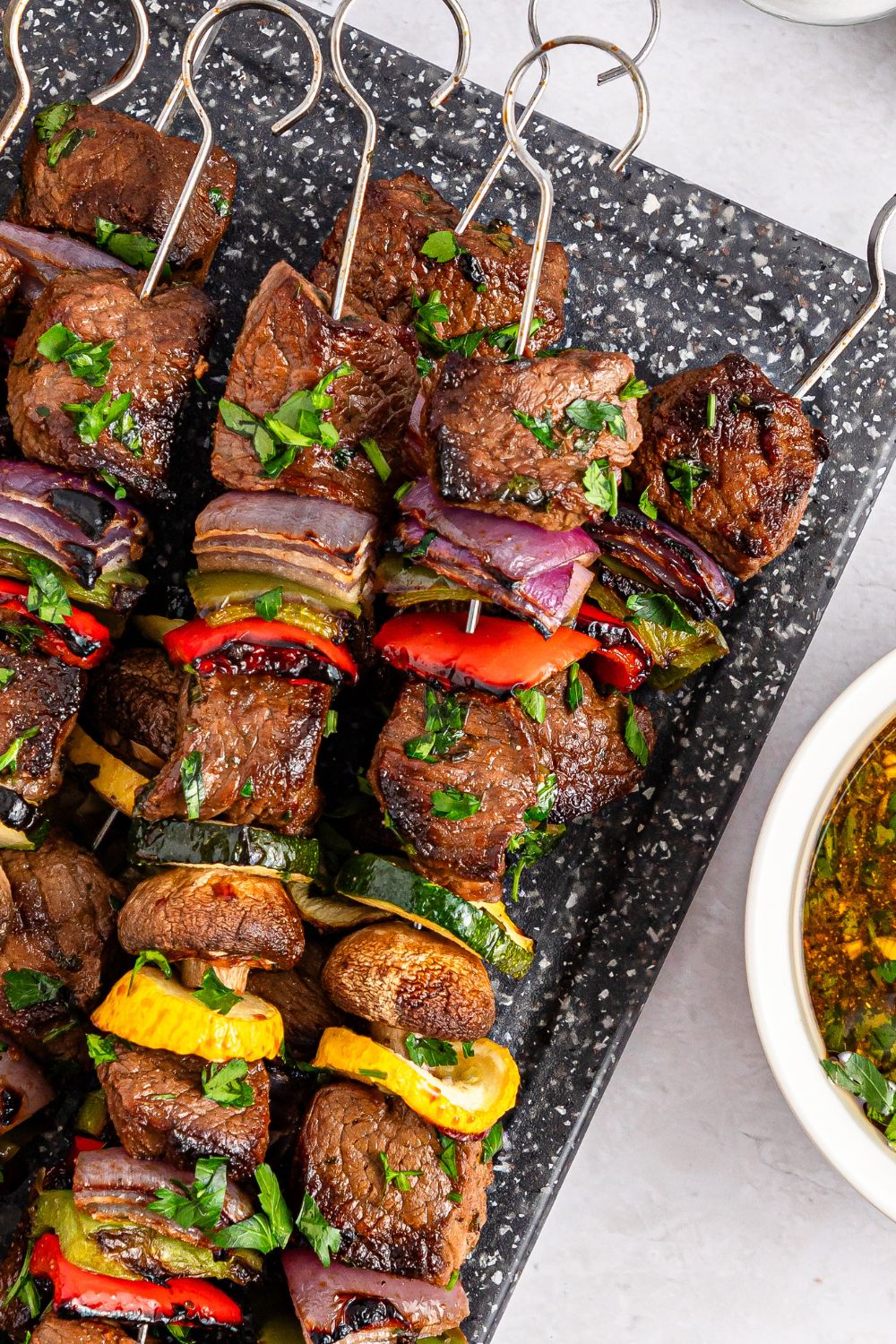 beef skewers with vegetables sitting on a tray with a bowl of extra marinade on the side