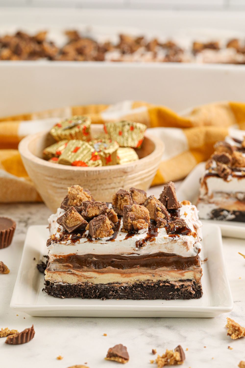 Reeses Lasagna sliced on table with a bowl of candies in background and pan behind it on table