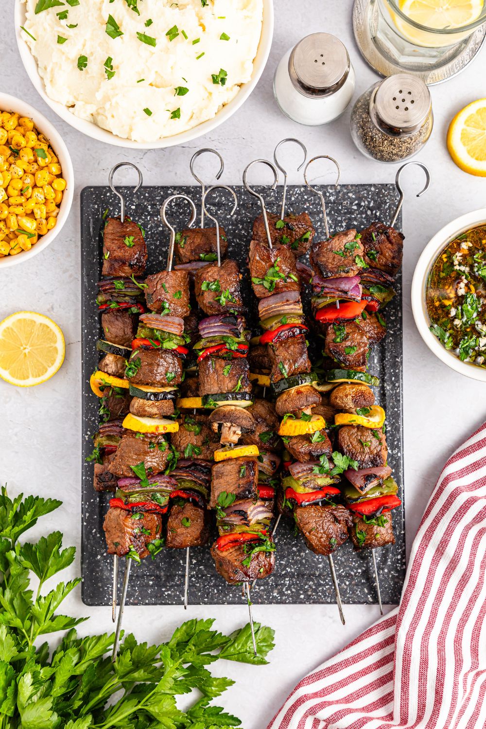 overhead look of Marinated Beef Kabobs on a tray with parsley, a bowl of corn, mashed potatoes and more all around the tray