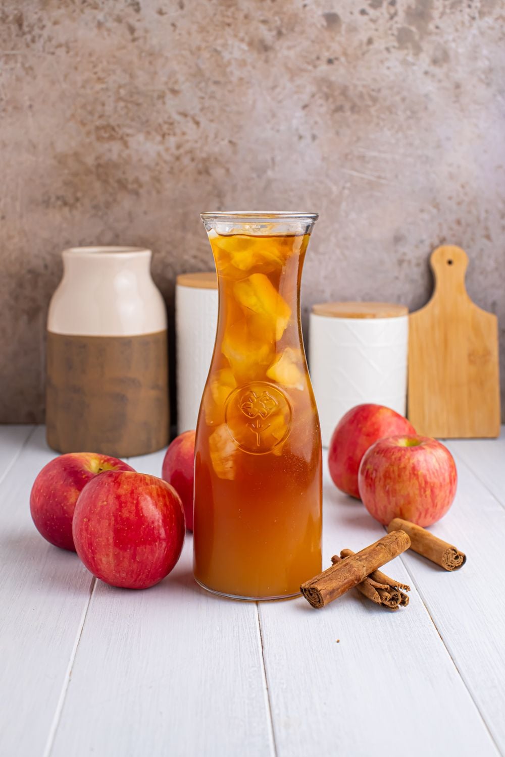 small pitcher of apple cider iced tea on table with apples and cinnamon sticks around it, and jars and cutting board in the background