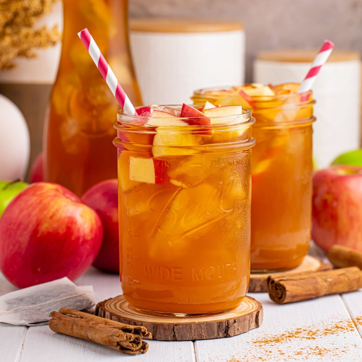 Apple Cider Iced Tea glasses on table with apples and cinnamon sticks around glasses and a red and white striped straw in each mason glass