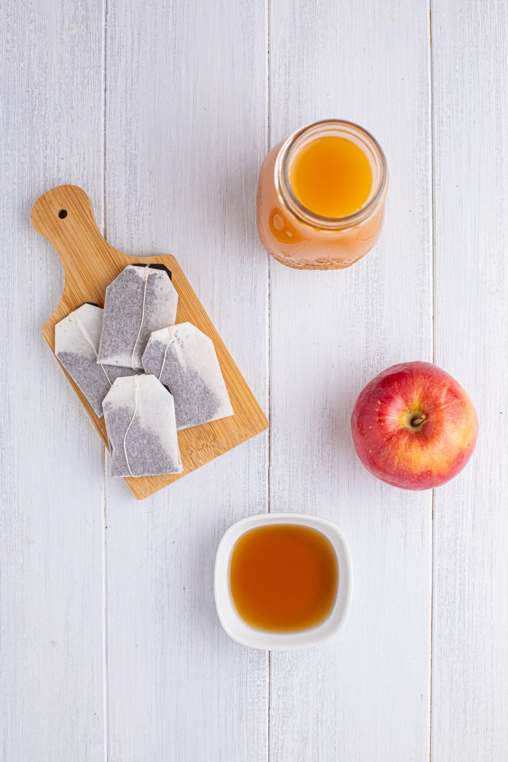 ingredients for making apple cider iced tea on a white table in single serve bowls: tea bags on a cutting board, mug of apple cider, whole apple