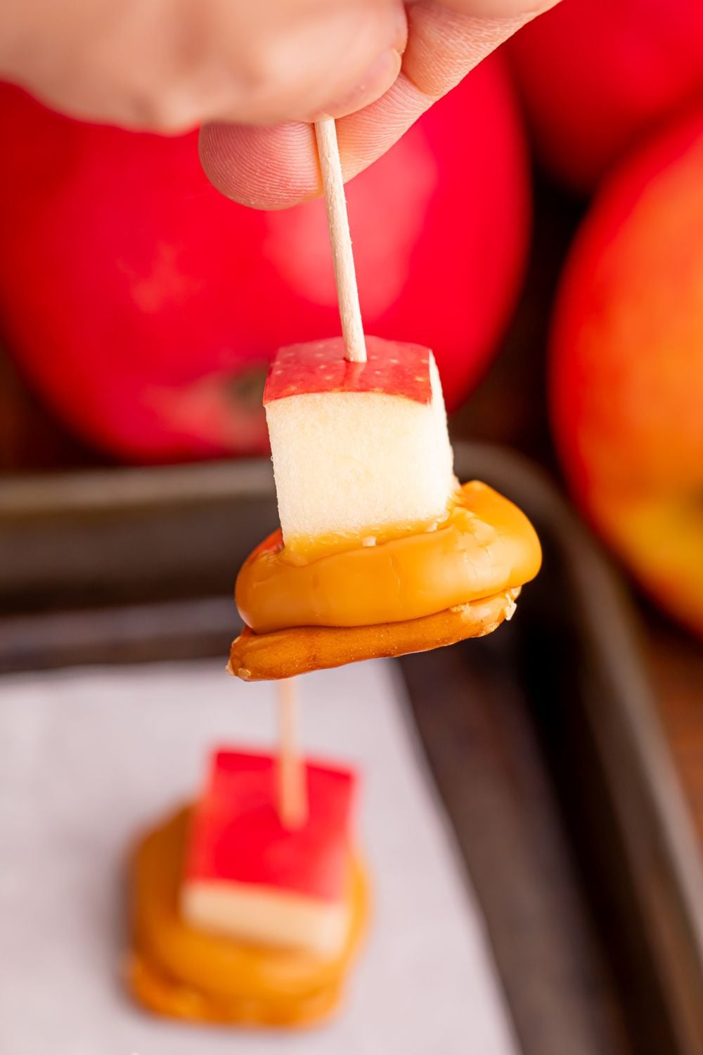 Caramel Apple Pretzel Bite being held in hand with toothpick with tray of more on table and apples around the tray