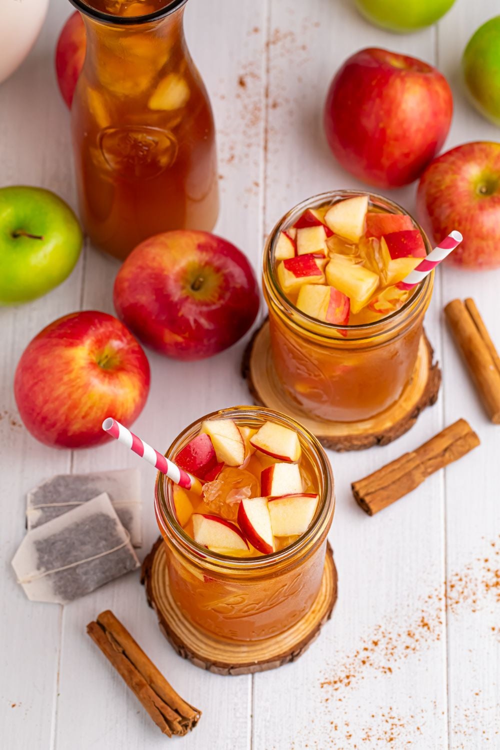 Apple Cider Iced Teas on table in two glasses, a large pitcher, with tea bags, cinnamon sticks and apples all over table