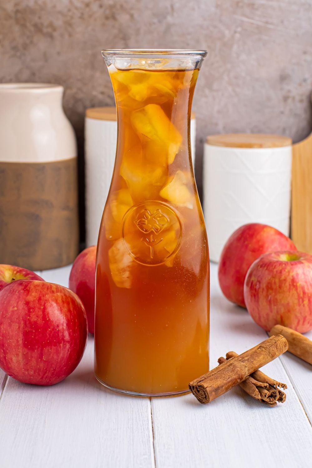 cider tea in a pitcher with cinnamon sticks and whole apples around it showing this is an apple cider iced tea