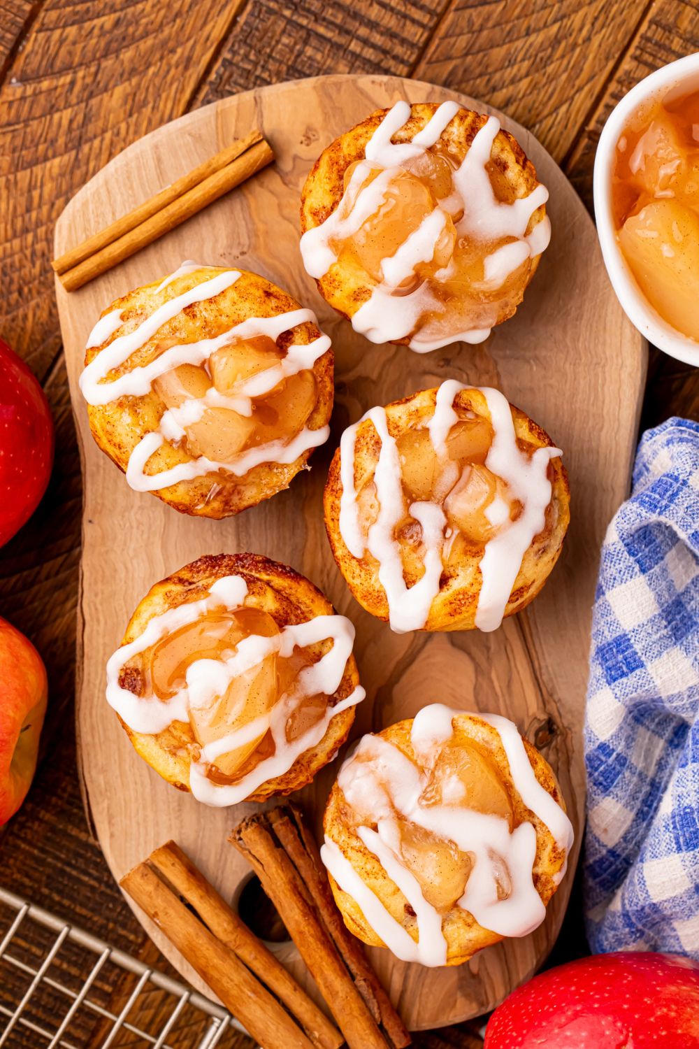 apple pie cuts placed on a wooden tray with cinnamon sticks around the cups, and a bowl of pie filling and towel to the side