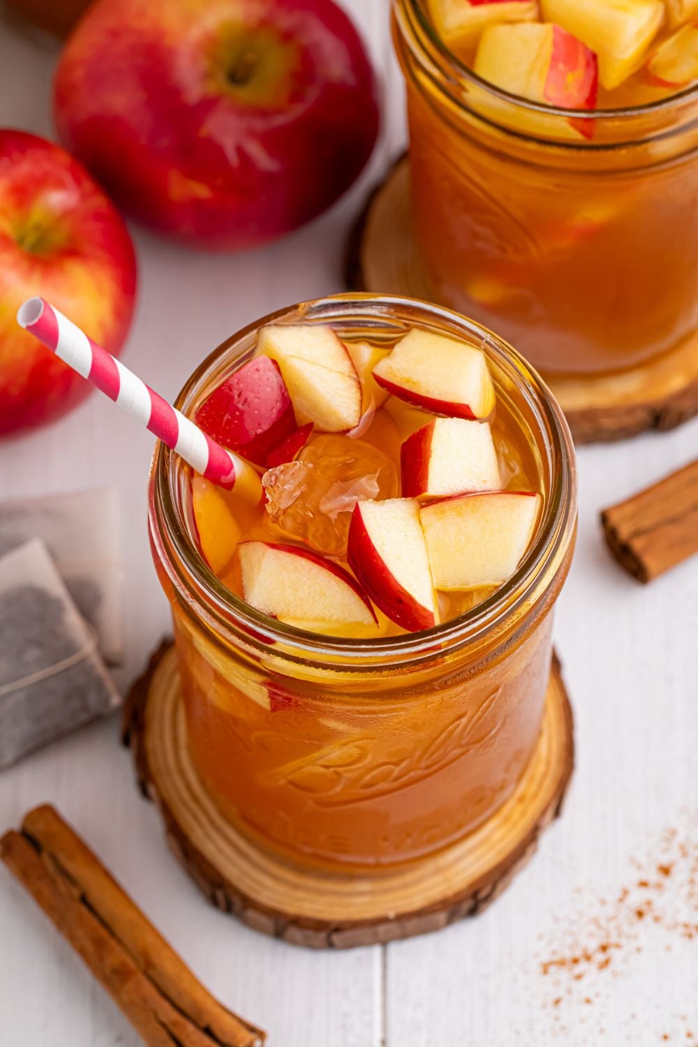 mason jar with an apple tea sitting on it on a wooden coaster and other glasses and apples spread over the table with a sprinkle of cinnamon on table