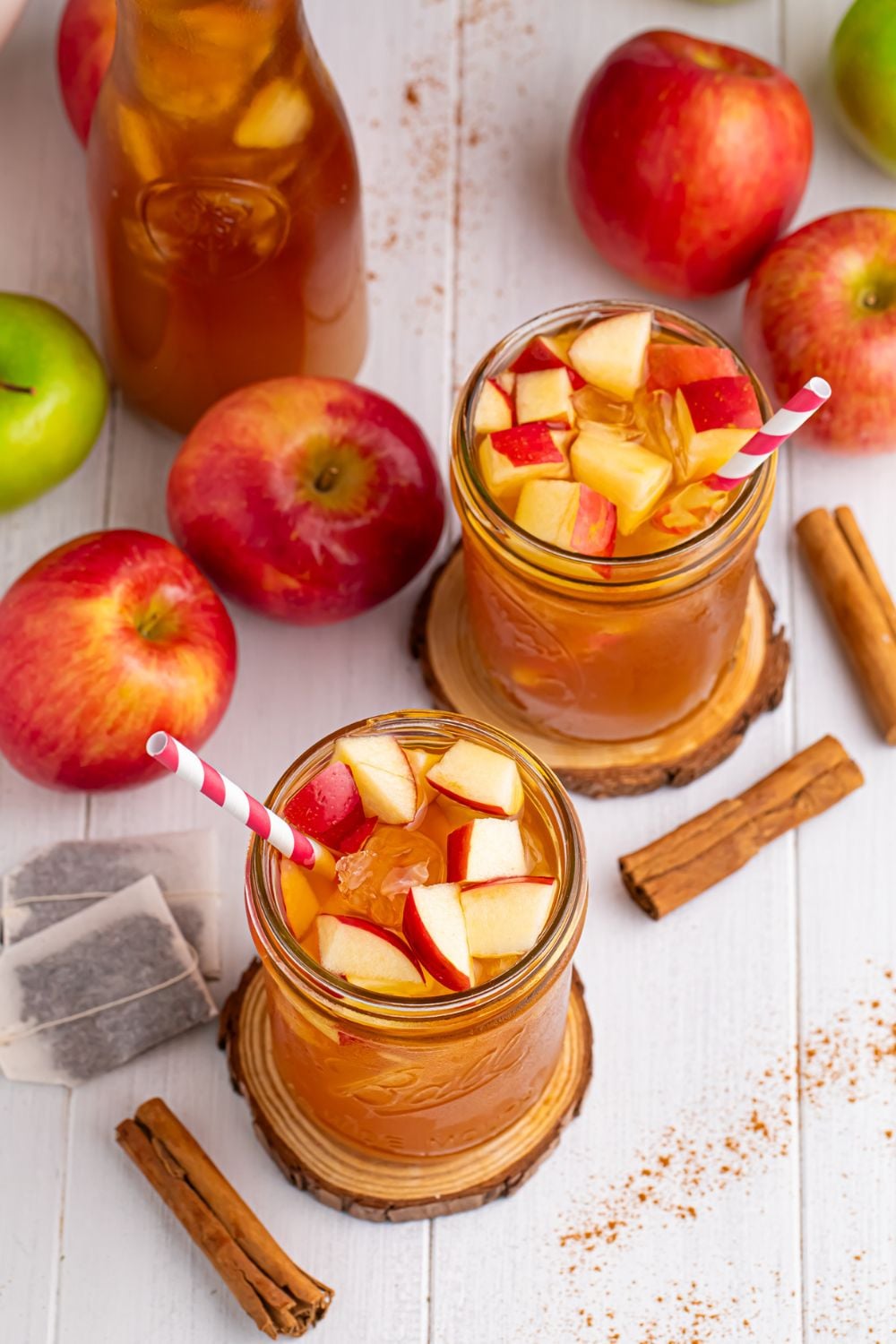 table setting with apple cider teas in glasses with straws. Tea bags, a variety of apples, cinnamon sticks and pitcher of tea all on table