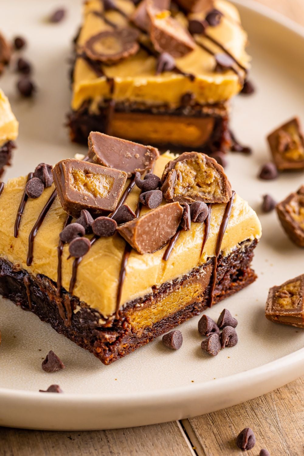 close up of a frosting Reeses Stuffed Brownie on table with mini chocolate chips and chopped peanut butter cups spread over table