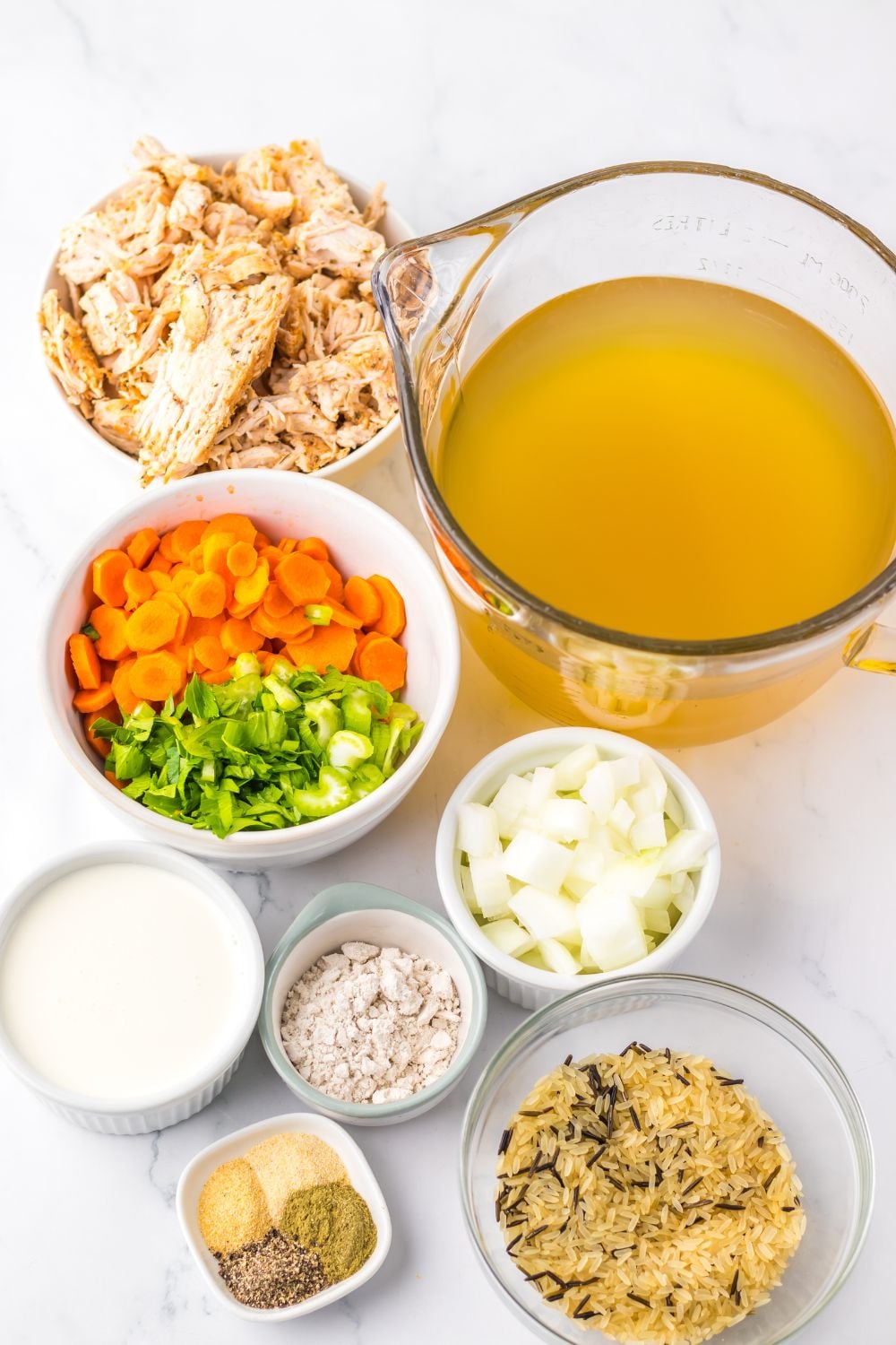 ingredients for this turkey and rice soup all in single bowls on table: stock, vegetables, diced turkey, seasonings, heavy cream, and more.