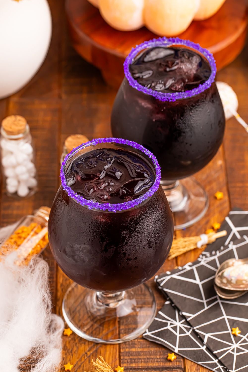 two glasses of witches brew cocktails sitting on wooden table with bottles of sprinkles in them, spider web napkins and more decor on table