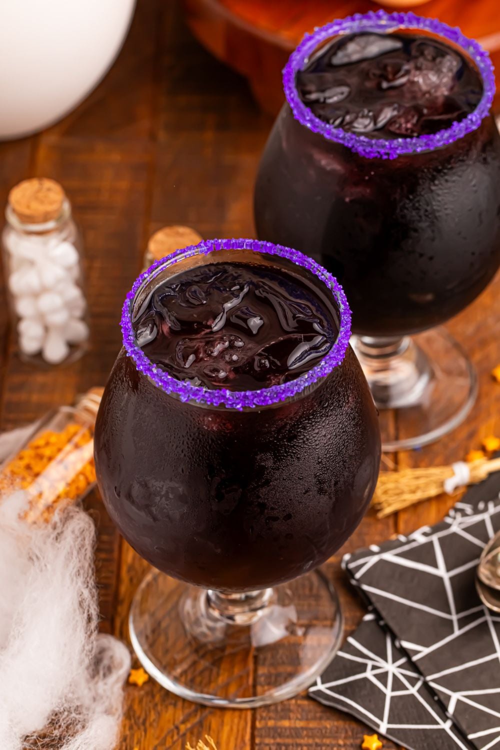 Witches Brew Cocktail on table with two glasses, a purple sugar rim and decor for Halloween all around it