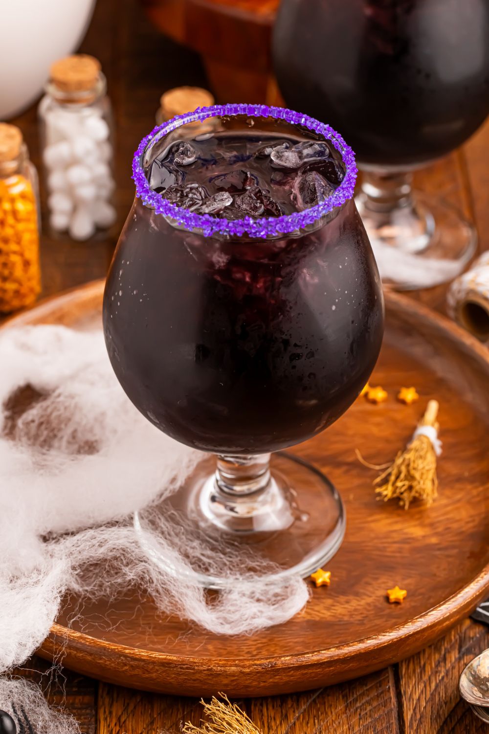 view of a glass with a deep purple cocktail in it with a light purple sugar rim sitting on table with Halloween decorations