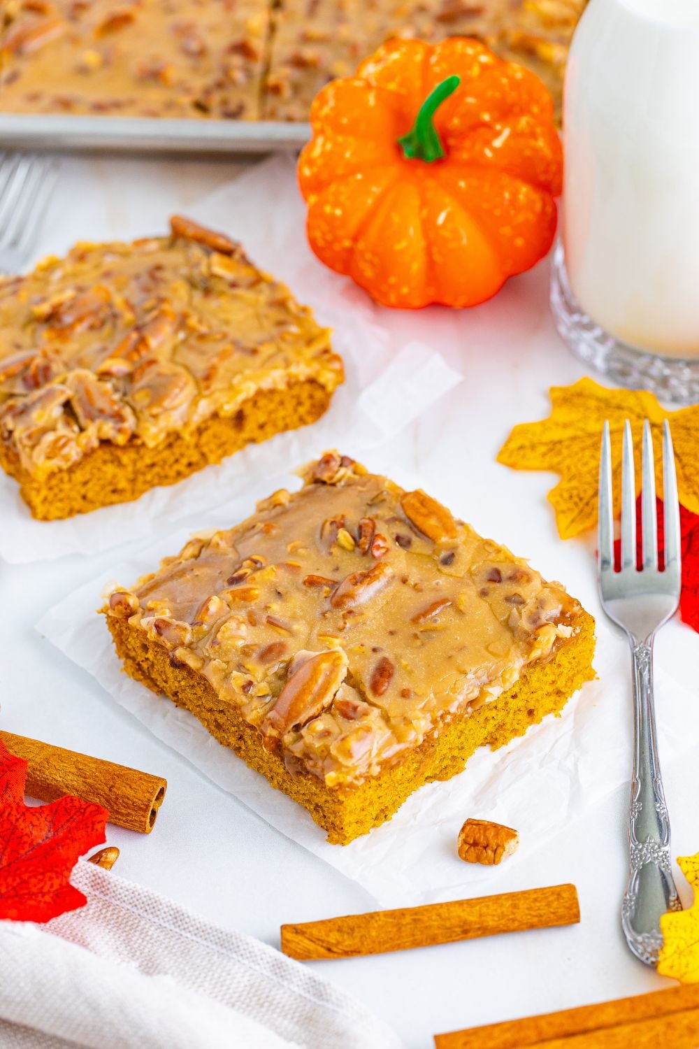 Pumpkin Snack Cake with Praline Icing sliced into squares and placed on table with a fork and faux pumpkin and glass of milk around the slices of cake