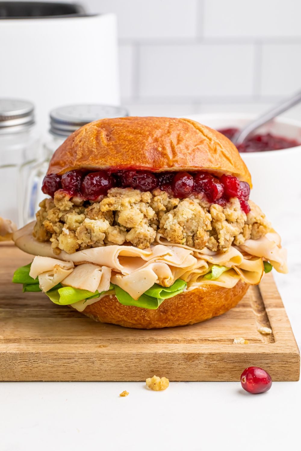 wooden cutting board on table with a turkey sandwich sitting on it with some crumbs and cranberries scattered on table