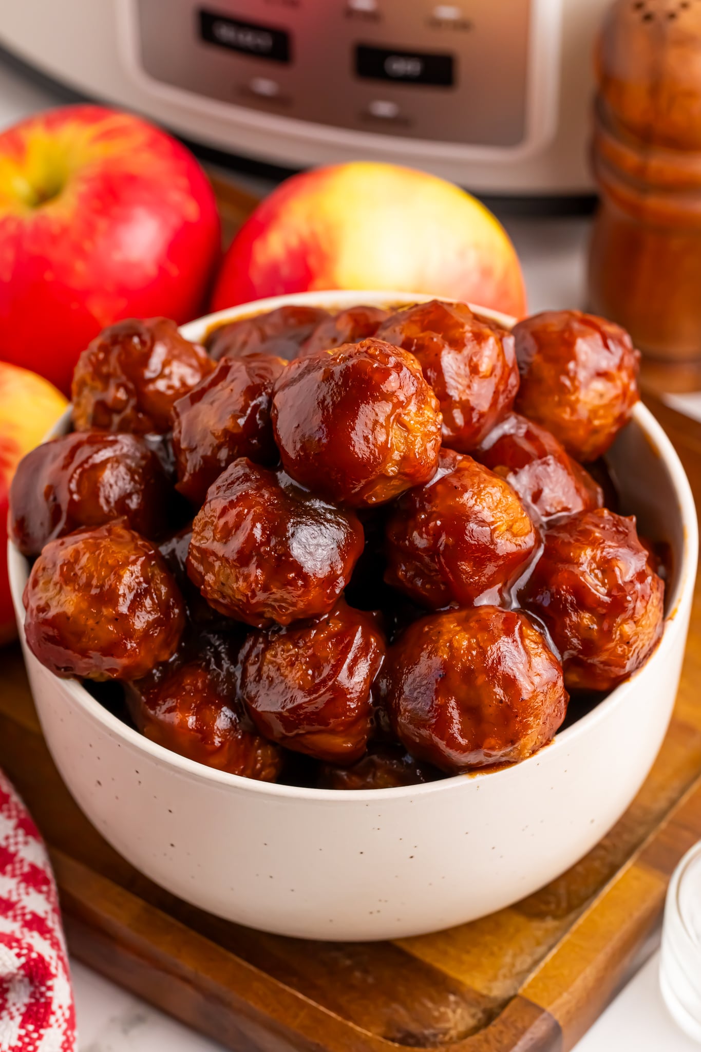 apple jelly meatballs in a bowl on table with slow cooker behind it on table and apples behind the bowl