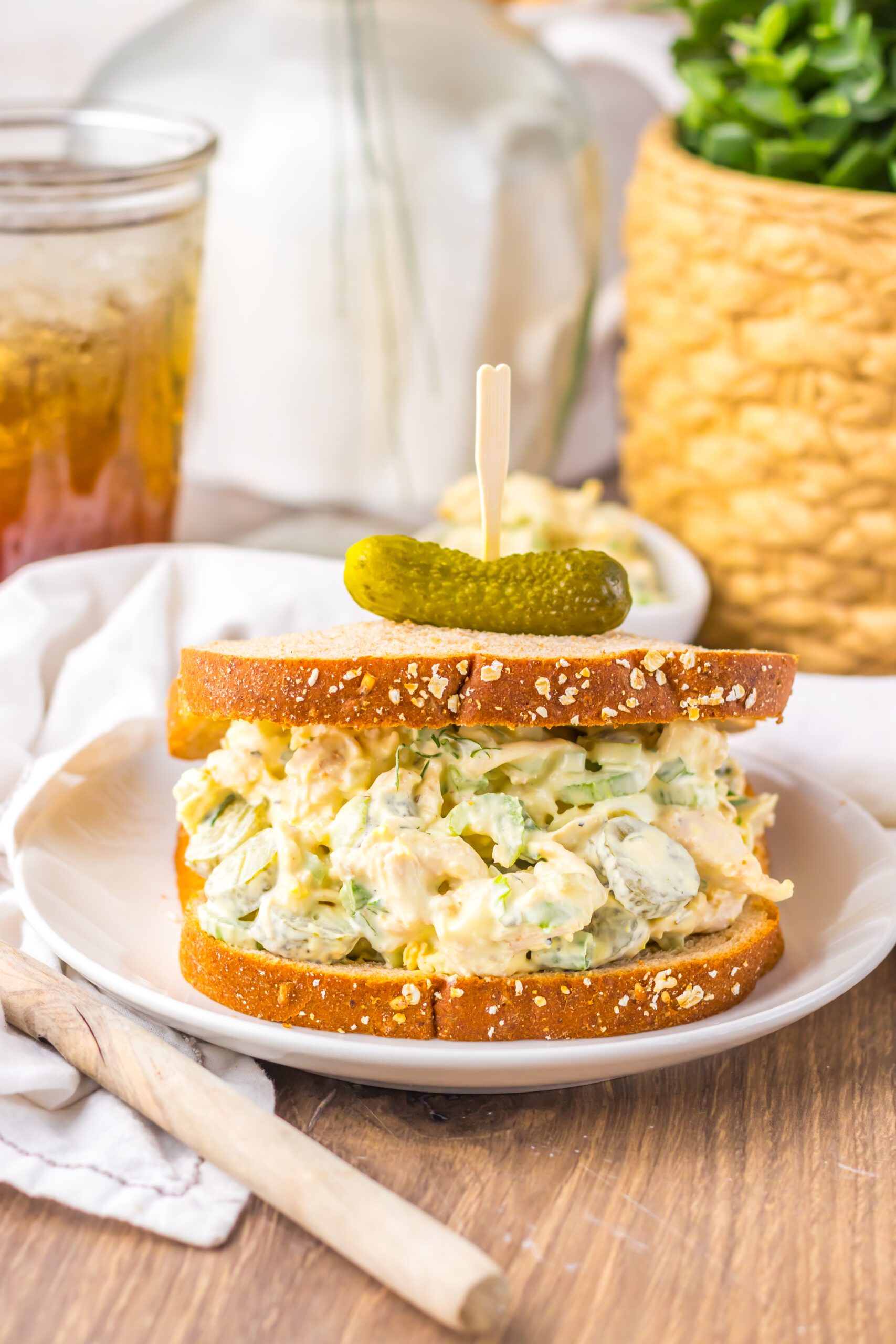 Pickle Chicken Salad sandwich on a white plate topped with a tiny pickle and iced tea in the background