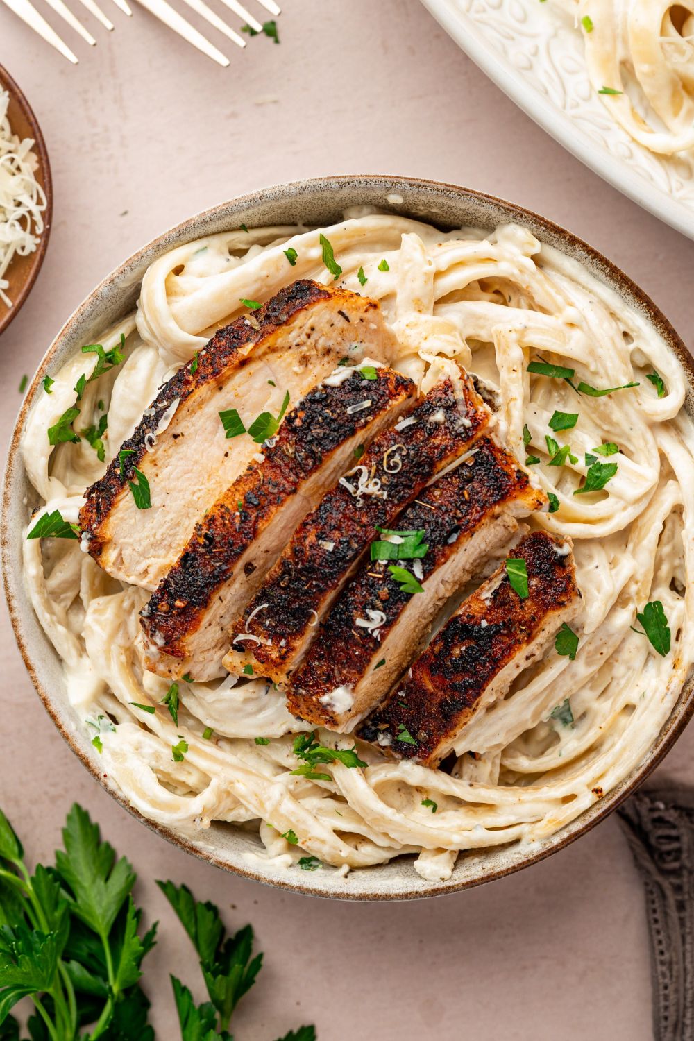 Blackened Chicken Alfredo in a bowl on table with parsley on the side, forks, and other items around the bowl 
