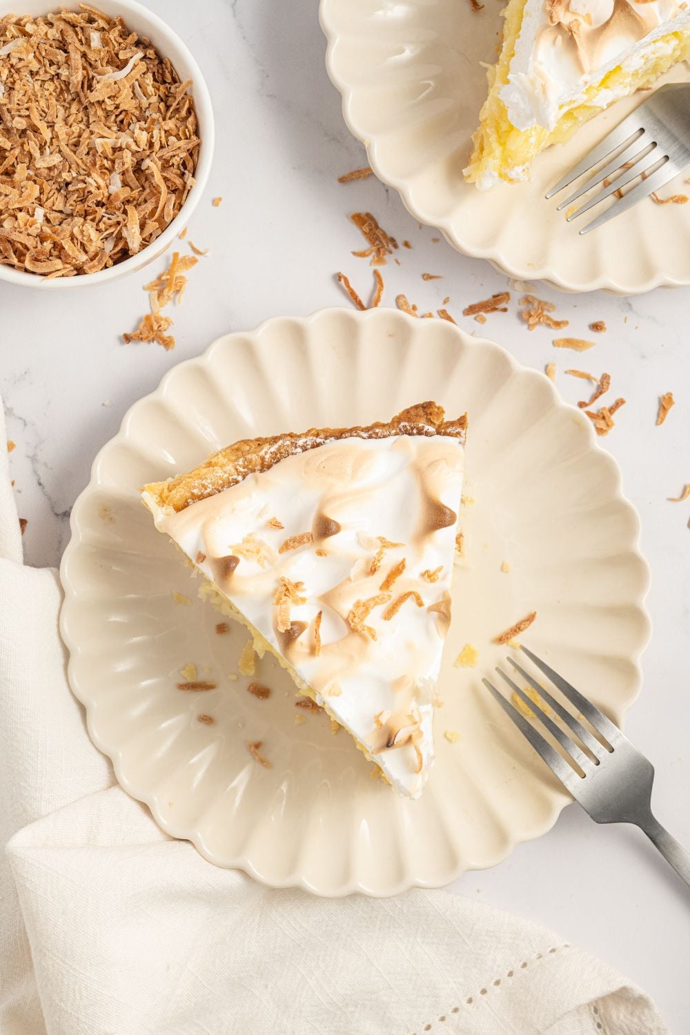 slices of Coconut Meringue Pie on plates with forks and toasted coconut flakes around the plate 