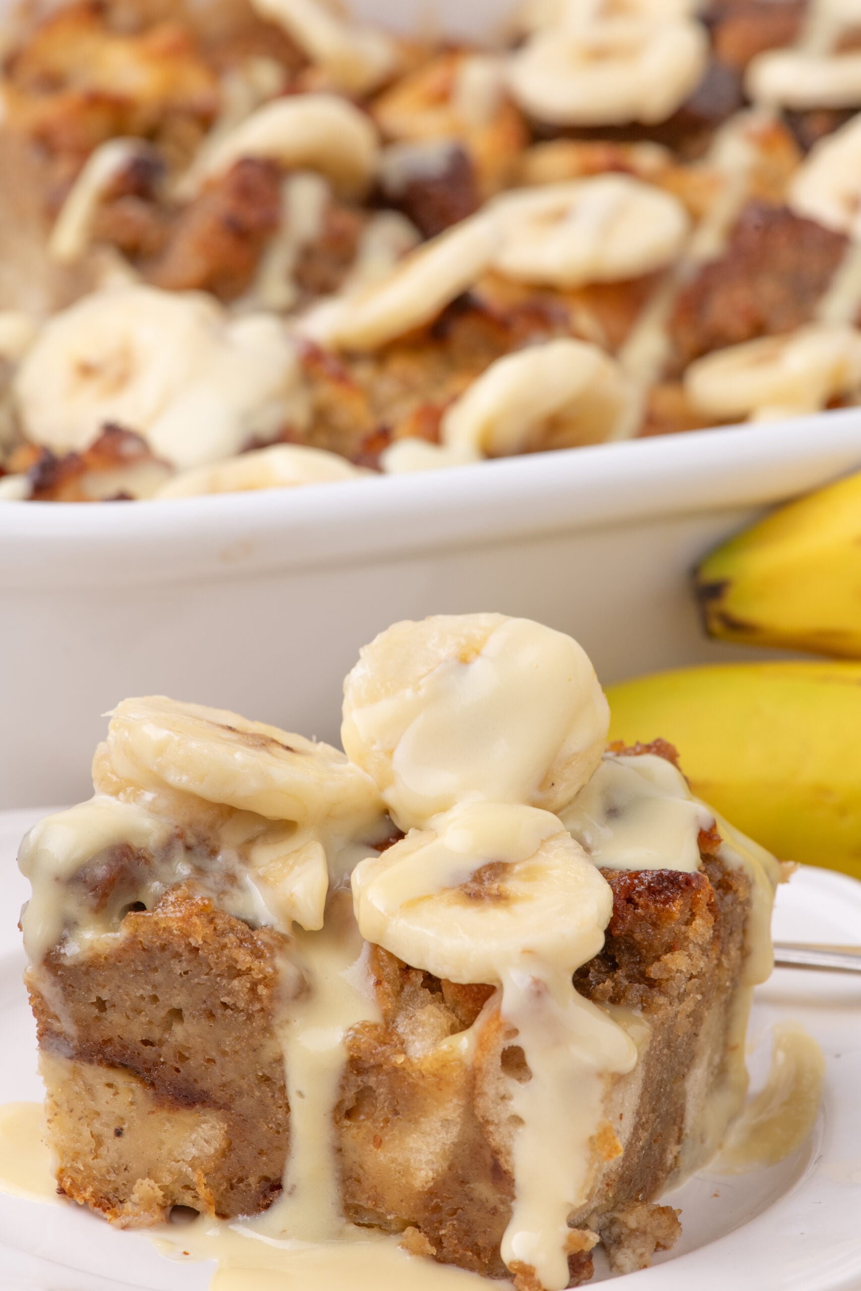 a white plate with a slice of Banana Bread Pudding on it topped with sliced bananas