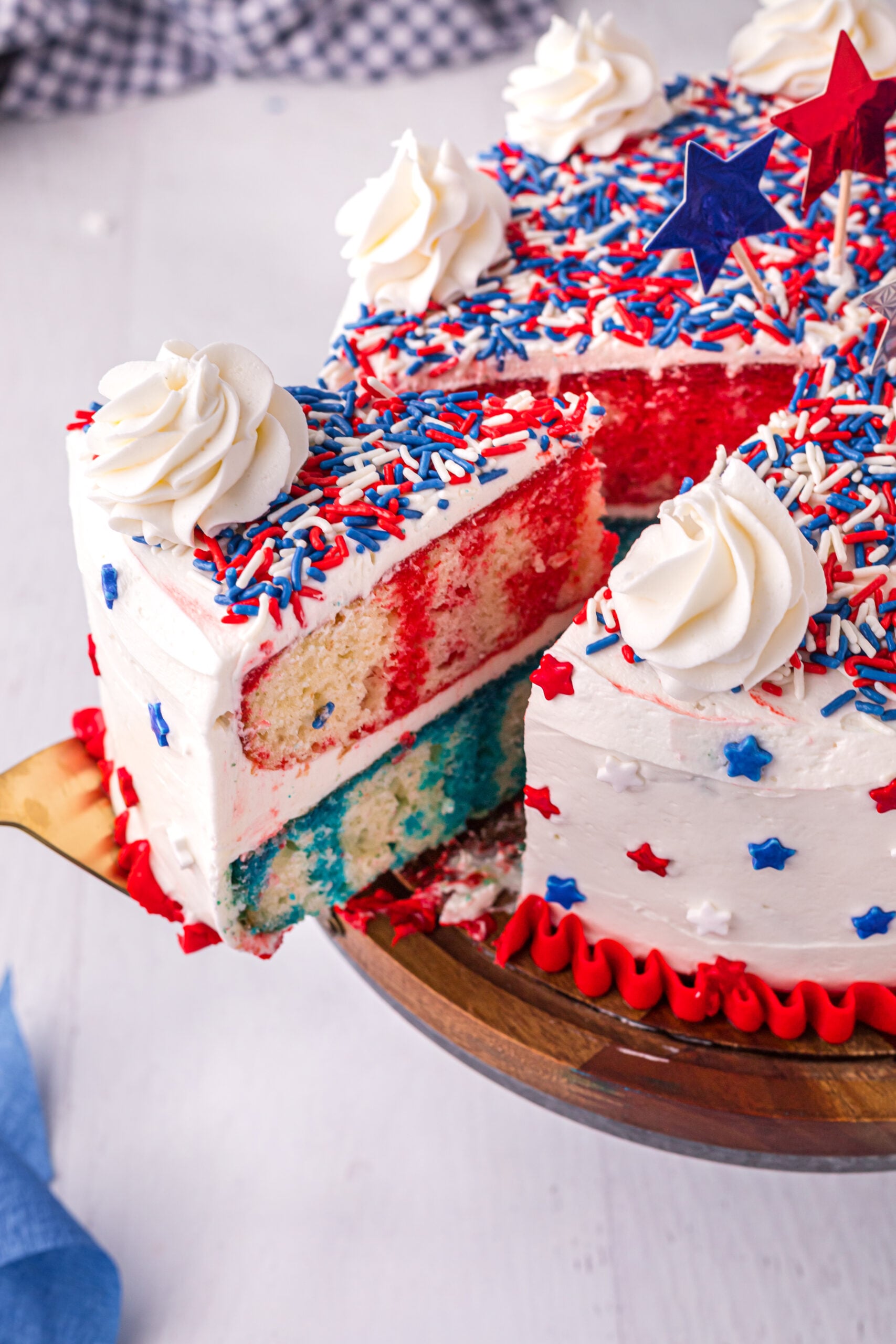 Side view of 4th of July Cake on a wood cake stand with a slice being removed by a gold cake server.
