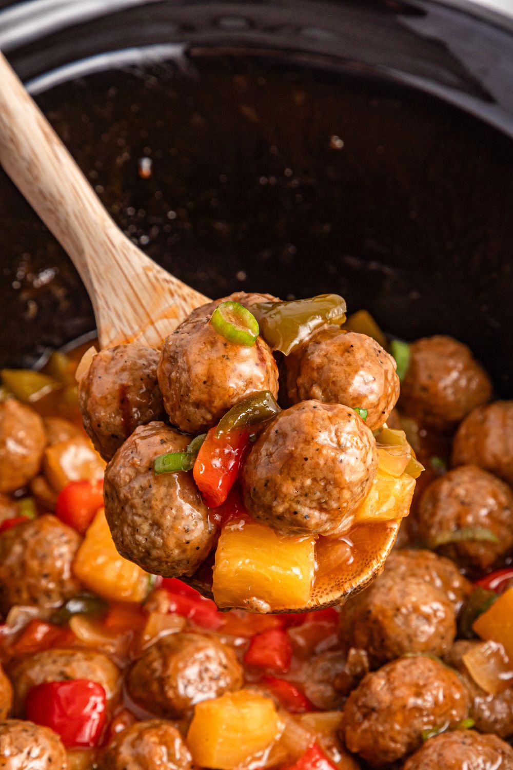 wooden spoon above crock pot with sweet and sour meatballs on spoon