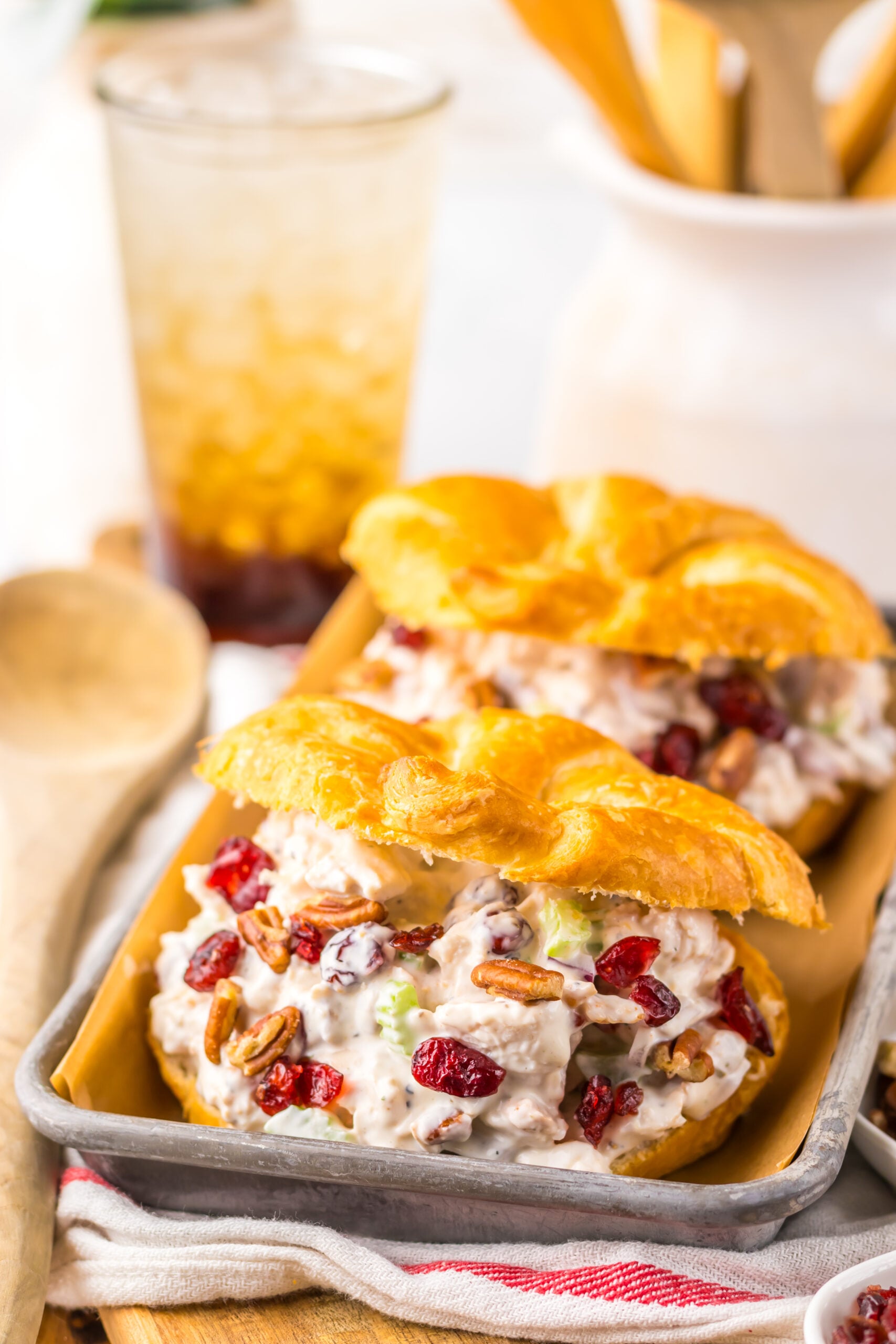 side view of a Cranberry Pecan Chicken Salad Croissant on a metal tray sitting with a glass of tea in the background