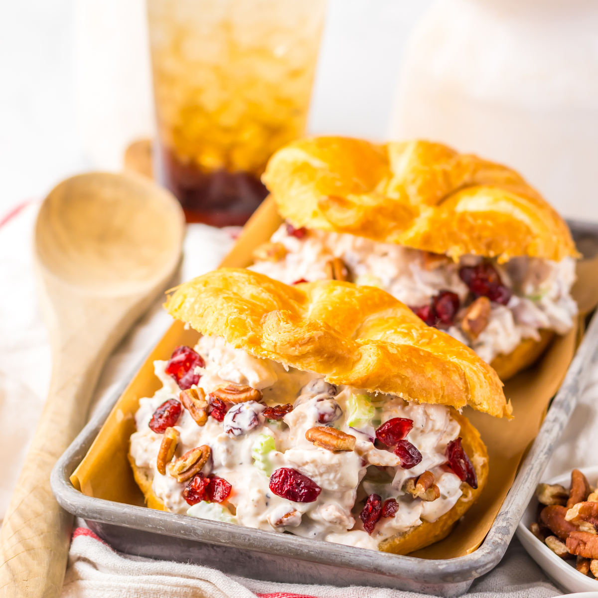 Close up view of a Cranberry Pecan Chicken Salad Croissant on a metal tray sitting on a red and white towel with pecans and a wooden spoon tot he side.