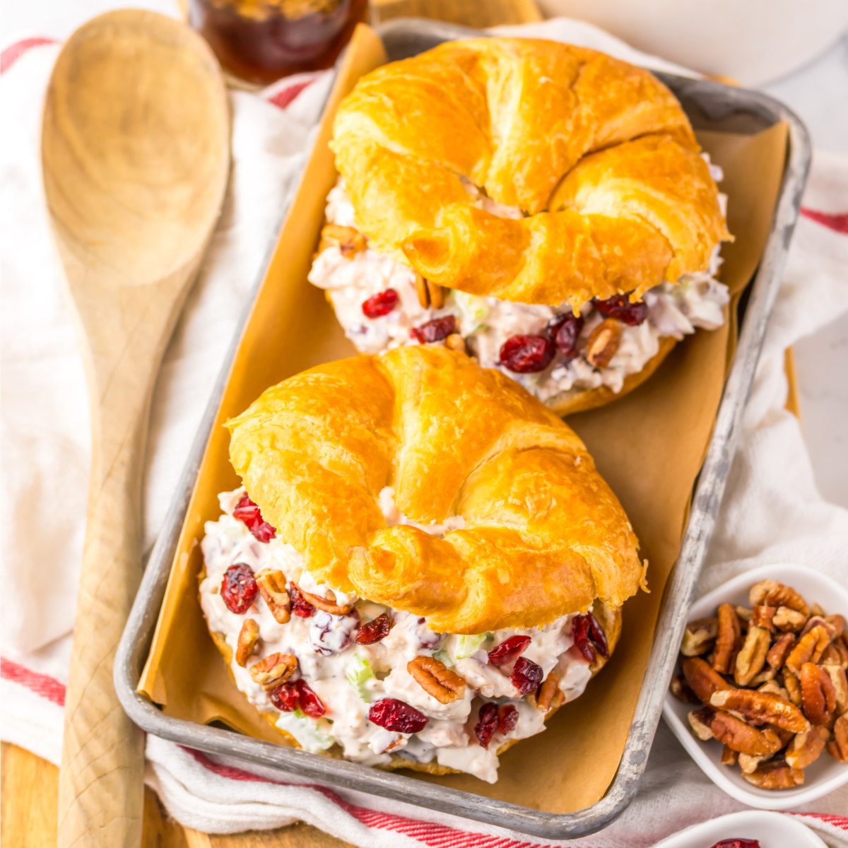 Over head view of a Cranberry Pecan Chicken Salad Croissant on a metal tray sitting on a red and white towel with pecans and a wooden spoon to the side.