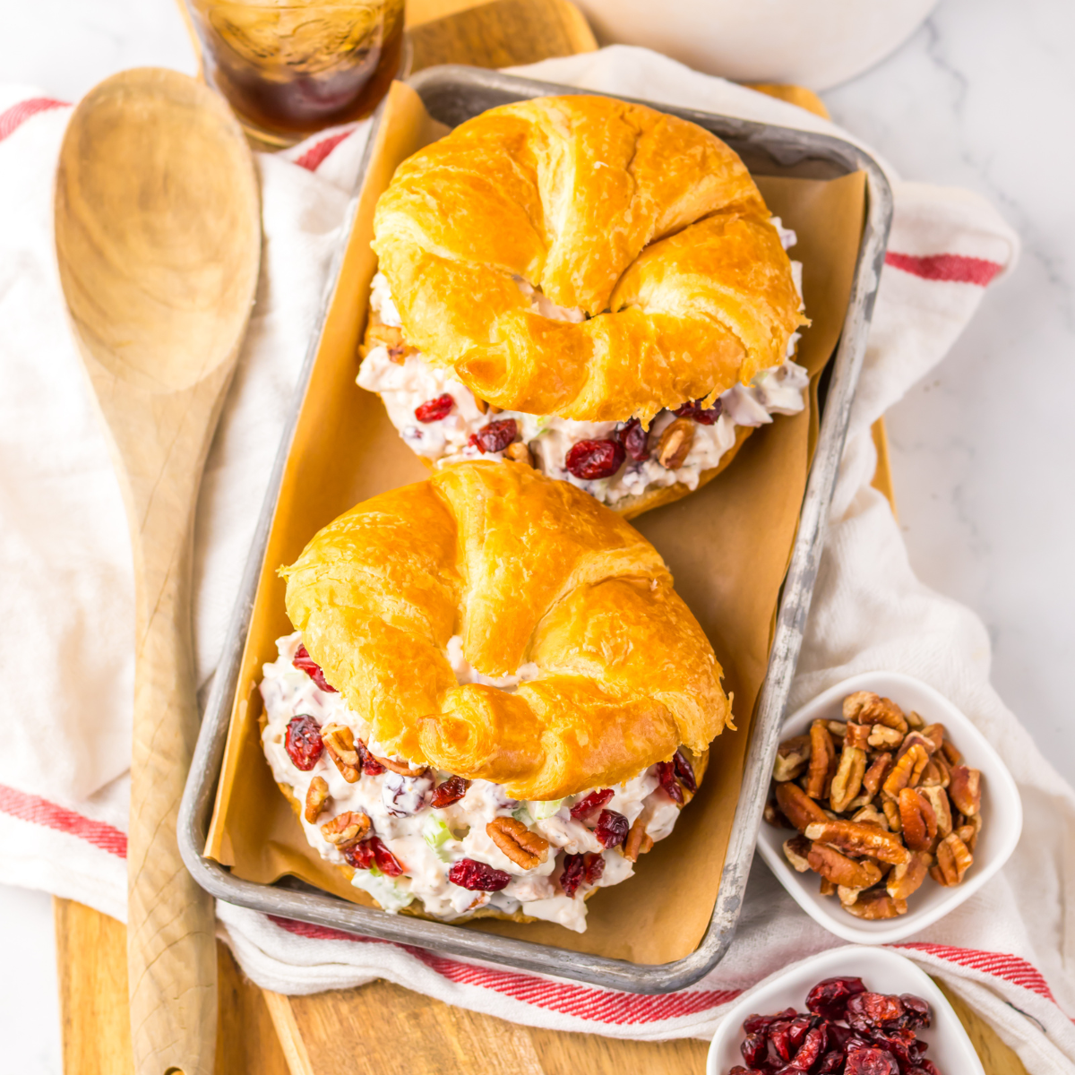 Close up view of a Cranberry Pecan Chicken Salad Croissant on a metal tray sitting on a red and white towel with pecans and a wooden spoon to the side.
